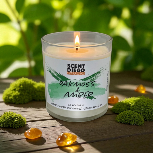 Candle labeled 'Oakmoss & Amber' from Scent diego Studio on a wooden surface with greenery in the background. There is pieces of Amber next to the candle. The candle wick is lit with a flame.