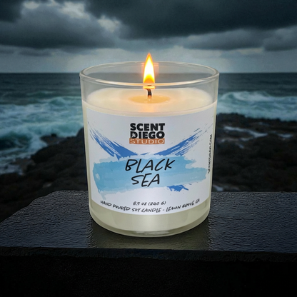 Candle labeled 'Black Sea' by Scented Diego Studio with ocean view in the background.  The wick is lit with a flame.