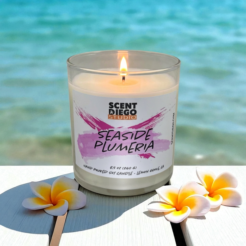Candle with 'Scented Diego Studio' label and plumeria flowers on a deck with the ocean in the background. 