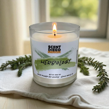 Candle with 'Scent Diego Studio' label on a white cloth with greenery.  There is no lid and a flame.