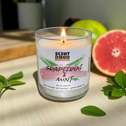 Grapefruit and mint candle with a label on a wooden surface with a half-cut grapefruit and leaves.  The wick is lit with a flame