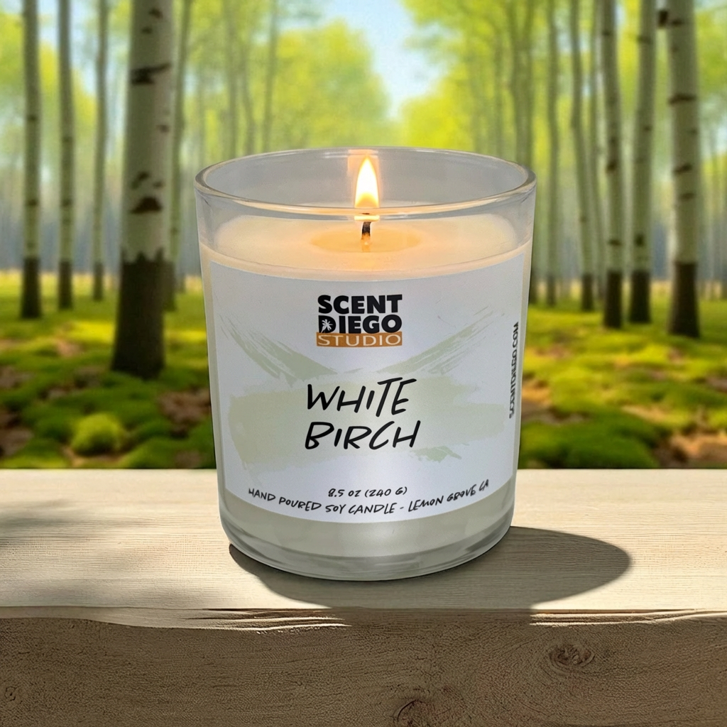Scented candle labeled 'White Birch' by Scent Diego Studio with a forest background.  The wick is lit with a flame.