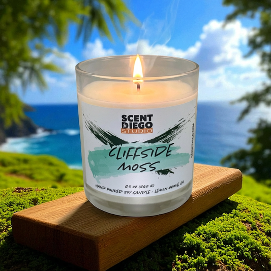 Candle labeled 'Cliffside Moss' by Scented Diego Studio with a scenic ocean background.  The wick is lit with a flame.