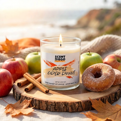Scented candle labeled 'Apple Cider Donut' surrounded by apples, a donut, and cinnamon sticks on a wooden surface with a blurred natural background. The candle is lit with a flame.